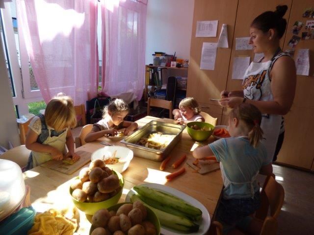 Der Blick richtet sich auf einen Tisch, auf dem Lebensmittel ausgebreitet sind. Um den Tisch herum sitzen vier Kinder, die auf Schneidebrettern Lebensmittel bearbeiten. Im rechten Hintergrund steht eine Erzieherin.