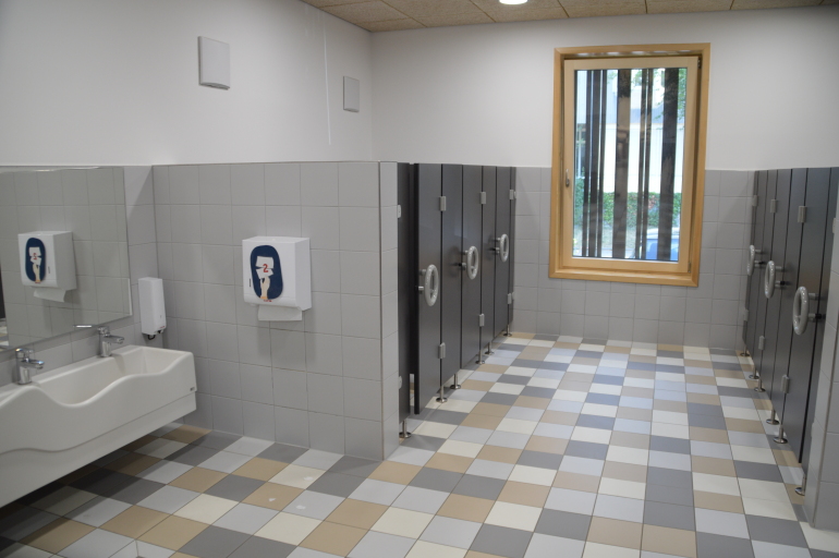 Ein Blick in den gefliesten Raum offenbart auf der linken Seite ein Kinderwaschbecken. Auf der rechten Seite im Hintergrund sind Türen zu erkennen, hinter denen sich Kindertoiletten befinden.
