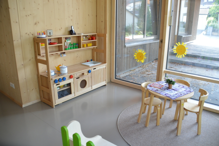 Blick in einen Raum mit großer Fensterfläche. Recht ein Kindertisch mit Stühlen, links an der Wand steht eine Kinderküche. 