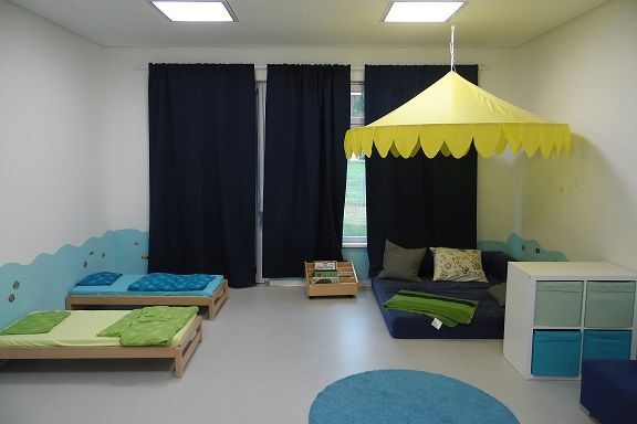 Blick in einen Raum, in dem sich links zwei Kinderbetten befinden. Rechts hängt ein Baldachin, unter dem eine Matratze mit vielen bunten Kissen liegt. Davor steht ein Regal.