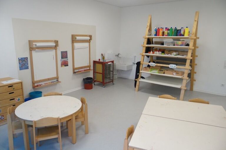 Blick ins Atelier, links befinden sich Wandstaffeleien und ein Materialschrank, rechts ein Materialregal, und im Vordergrund stehen Tische.