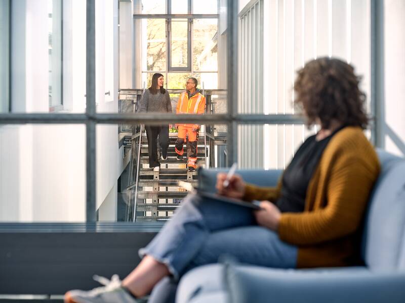 Im Vordergrund sitzt eine Frau. Im Hintergrund laufen zwei Personen eine Treppe herab und sprechen miteinander.