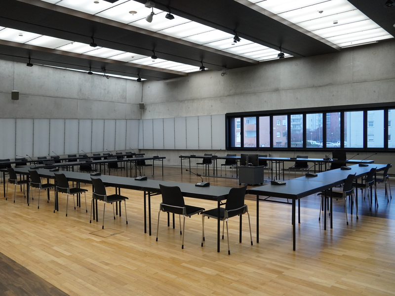 Ein großer, moderner Konferenzraum mit langen Tischen und Stühlen um eine zentrale Anordnung, Fenstern an der rechten Seite und einer hellen Deckenbeleuchtung.