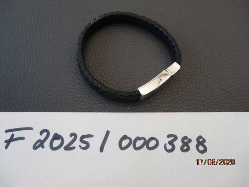 Ein schwarzes Armband mit Metallschließe liegt auf einer dunklen Oberfläche, daneben eine handgeschriebene Nummer und ein Datum. KI-BildBeschreiber