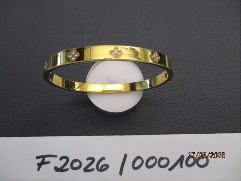 Ein glänzender goldener Armreif mit kleinen verzierten Blumen liegt auf einer schwarzen Oberfläche, neben einer handgeschriebenen Zahlenfolge. KIBildBeschreiber