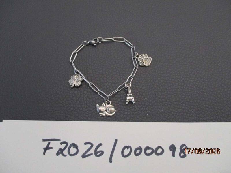 Ein Armband mit Kettengliedern und vier Anhängern (Kleeblatt, Katze, Eiffelturm, Pfote) liegt auf einer dunklen Oberfläche, darunter ein Zettel mit Zahlen und einem Datum.