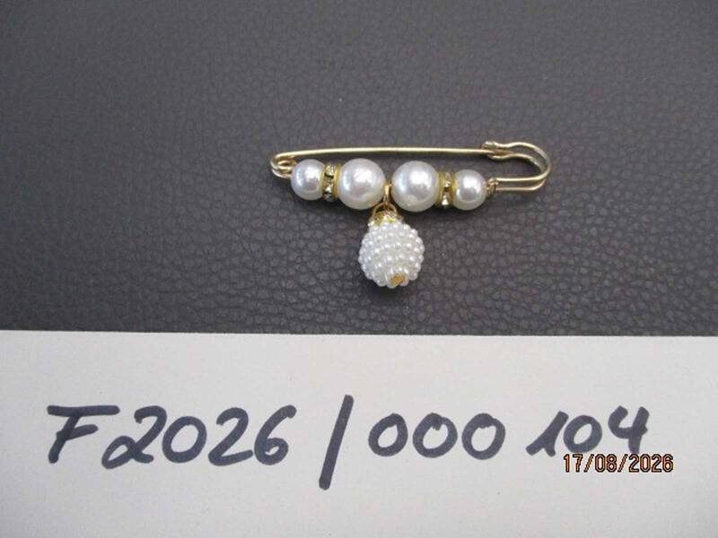 Eine goldfarbene Anstecknadel mit mehreren aufgereihten Perlen und einem herabhängenden runden Schmuckstück liegt auf einer dunklen Oberfläche, darunter handschriftlich "F2026 / 000 104". KI-BildBeschreiber