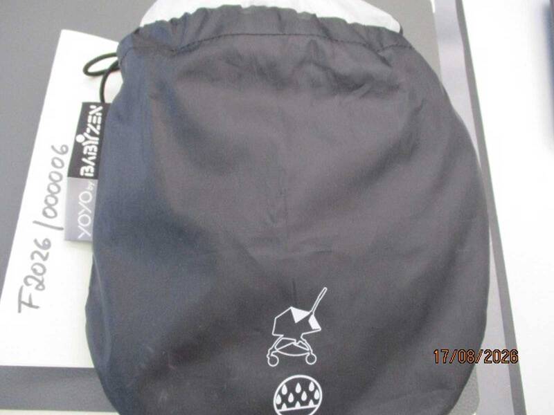 Ein schwarzer Kordelzugbeutel mit einem Symbol für einen Kinderwagen und Regentropfen darauf liegt auf einem weißen Papier mit der Aufschrift "BABYZEN YOYO" und einem Datum im unteren rechten Eck. KI-BildBeschreiber