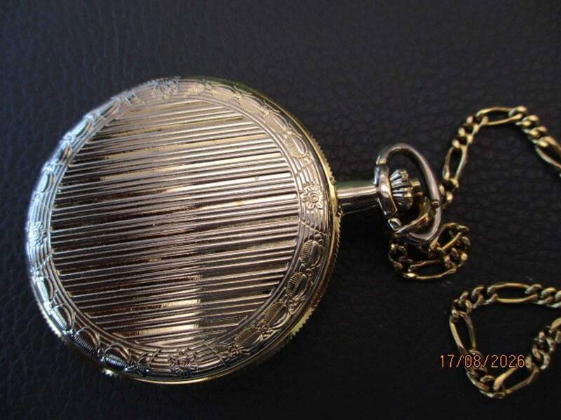 Eine goldene Taschenuhr mit glatter Oberfläche und Gravuren liegt auf einem dunklen Hintergrund, an einer goldenen Kette befestigt. KI-BildBeschreiber