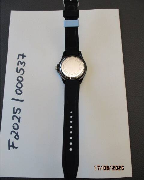 Die Rückseite einer Armbanduhr mit schwarzem Armband liegt auf einem weißen Papier, auf dem seitlich alphanumerische Zeichen notiert sind. KI-BildBeschreiber