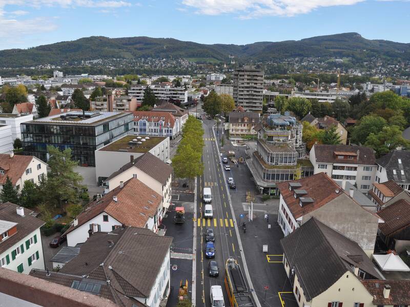 Das Luftbild zeigt eine breite Straße in einer Stadt. Im Hintergrund sind Berge zu sehen. 