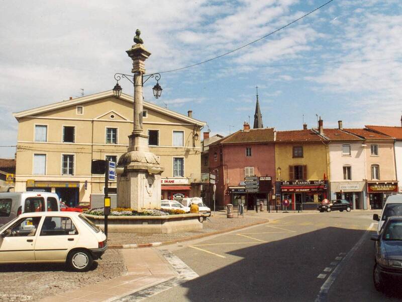 Zu sehen ist ein sonniger Marktplatz mit Brunnen, Autos und Cafés. 