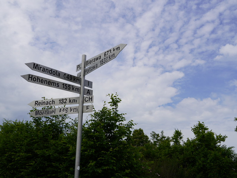 Ein Wegweiser zeigt in verschiedene Richtungen, mit Entfernungsangaben zu verschiedenen Städten, vor einem bewölkten Himmel.