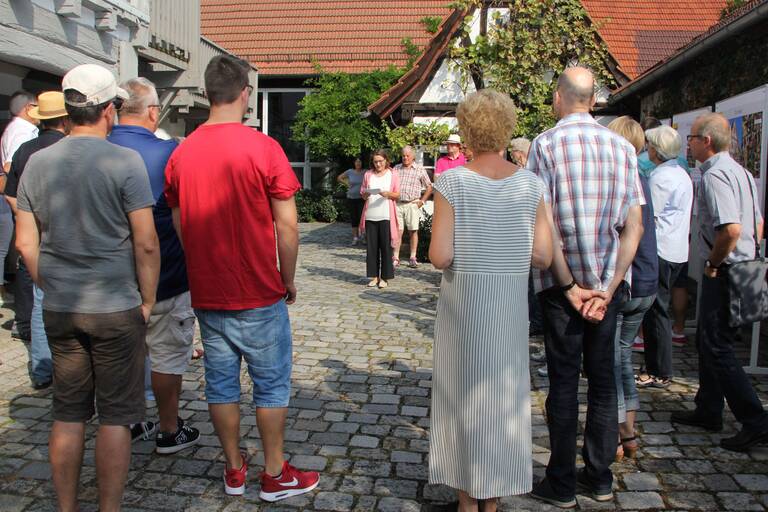 Eine Gruppe von Menschen steht auf einem gepflasterten Innenhof und hört einer Person zu, die vorne spricht. (KI-Bildbeschreiber)