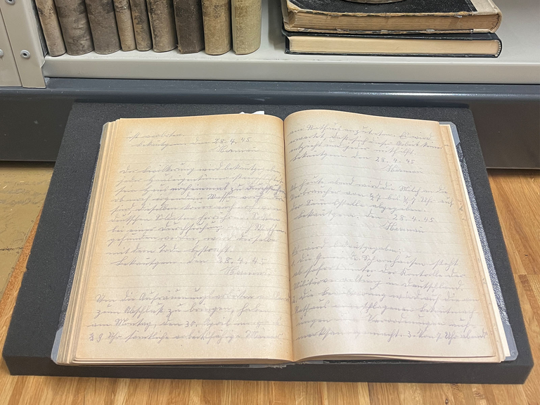Ausschellbuch Ein altes aufgeschlagenes, handgeschriebenes Buch, das Ausschellbuch, liegt auf einem Schaumstoffpolster, umgeben von alten, gebundenen Büchern im Regal.