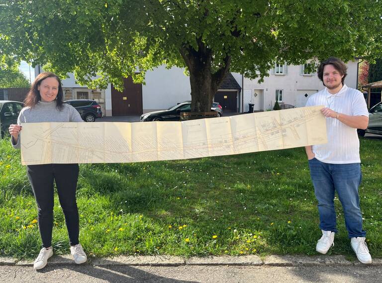 Lageplan der Esslinger Straße Zwei Personen stehen im Freien auf einer Wiese und halten gemeinsam einen langen, ausgebreiteten Plan in den Händen.