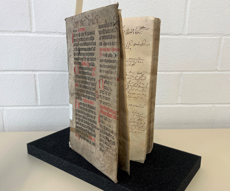 Bücher im Stadtarchiv Ein altes Buch steht geöffnet auf einem schwarzen Sockel, mit handschriftlichem Text und gedruckten, teils roten Buchstaben auf den Seiten.