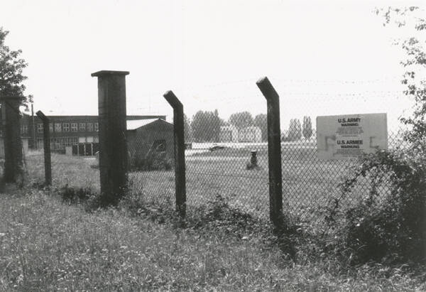 Ein eingezäuntes Areal mit einem Schild, das "U.S. Army" zeigt, dahinter Gebäude und Gras.