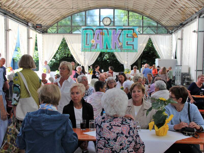 Mehrere Menschen sitzen an Biertischen in einer geschlossenen Halle, während ein großes Schild mit der Aufschrift "DANKE" im Hintergrund hängt.