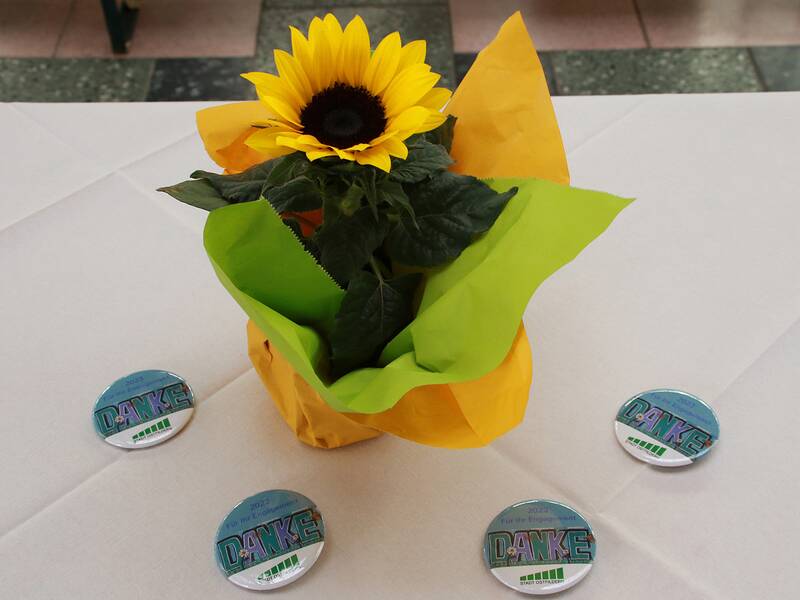 Eine Sonnenblume steht in einem gelb-grünen Papier eingewickelt auf einem Tisch, umgeben von vier "Danke"-Ansteckern.