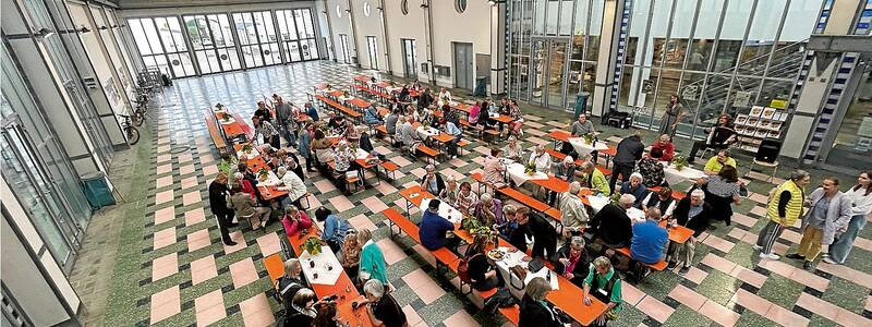 In einer großen, lichtdurchfluteten Halle sitzen viele Menschen an langen Tischen und unterhalten sich.