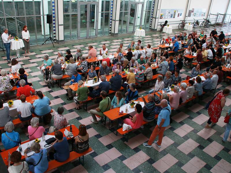 Eine große Gruppe von Menschen sitzt in einer Halle auf orangefarbenen Bänken und Tischen, während zwei Personen vorne stehen und sprechen.