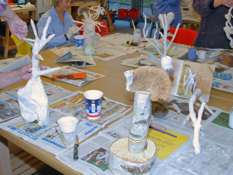 Auf dem Bild sind mehrere Menschen zu sehen, die an einem Tisch in einem Atelier arbeiten. Sie gestalten Skulpturen aus Holzstücken und Gips. Auf dem Tisch liegen Zeitungen, Werkzeuge und Behälter.
