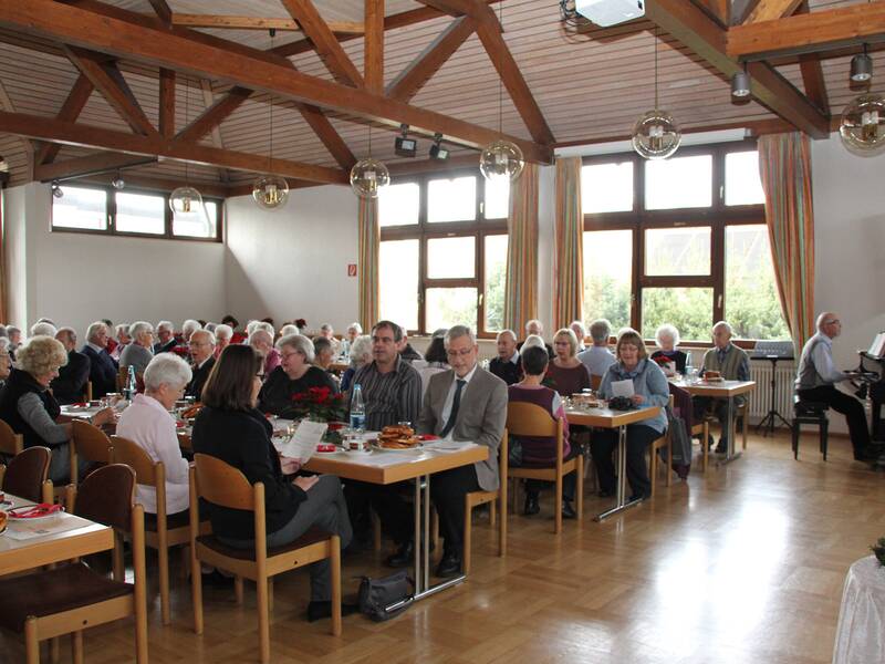 In einem Saal sitzen viele ältere Menschen an Tischen und unterhalten sich, während ein Mann Klavier spielt.