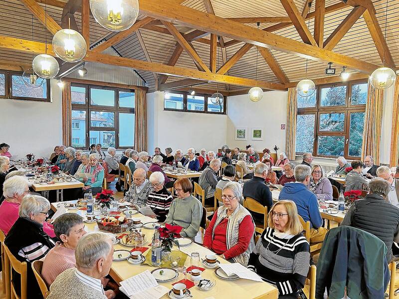 In einem holzgetäfelten Raum sitzt eine große Gruppe älterer Menschen an mehreren Tischen und unterhält sich, während sie gemeinsam Kaffee trinken und Kuchen essen.