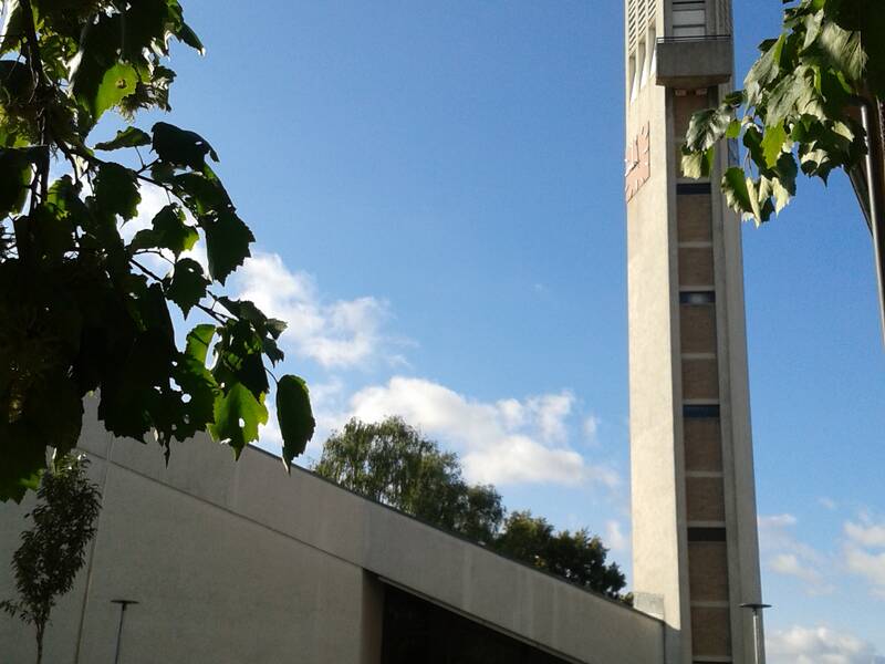 Die Aufnahme zeigt einen modernen Kirchturm und ein abgewinkeltes Kirchendach, eingerahmt von grünen Blättern, vor einem blauen Himmel.