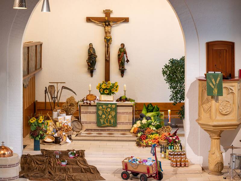 Ein Kirchenaltar, geschmückt mit Erntegaben, darunter Obst, Gemüse, Brot und Blumen, steht im Zentrum eines hellen Raumes mit einem Kruzifix und zwei Heiligenfiguren an der Wand.