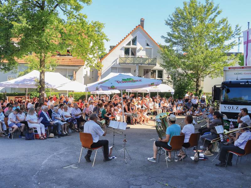 Zum Open-Air-Gottesdienst beim Gemeindefest spielt eine Blaskapelle im Freien, während viele Menschen in einem Halbkreis unter Pavillons und im Schatten von Bäumen das Konzert beobachten.
