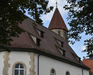 Die St. Blasius Kirche mit einem steilen, roten Ziegeldach und einem Turm mit einem goldenen Kreuz steht umgeben von Bäumen und Sträuchern.