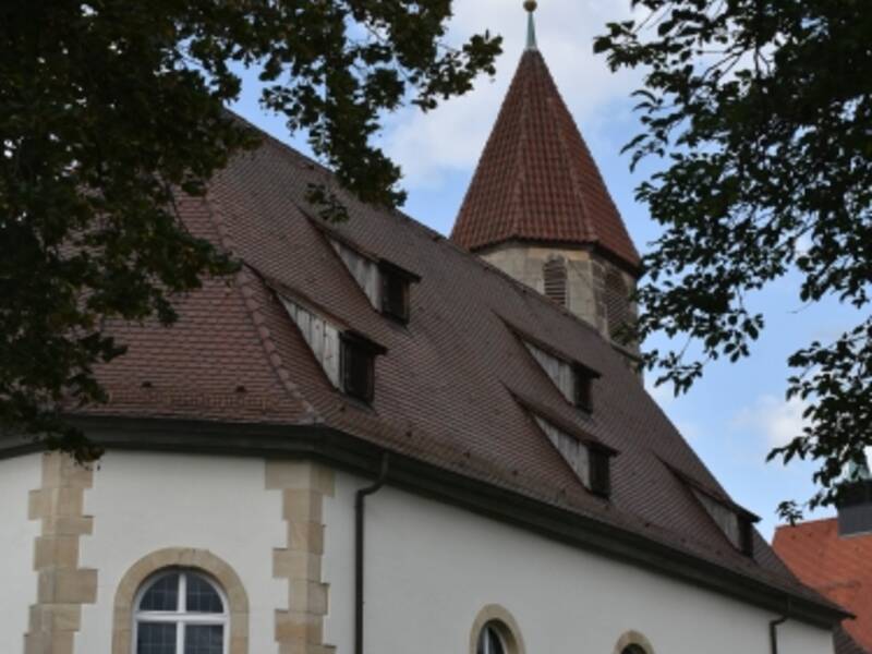 Die St. Blasius Kirche mit einem steilen, roten Ziegeldach und einem Turm mit einem goldenen Kreuz steht umgeben von Bäumen und Sträuchern.