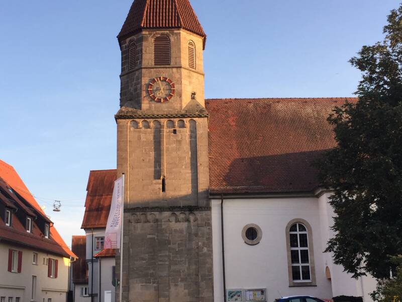 Die St. Blasius Kirche mit einem hohen, spitzen Turm und einer großen Uhr an der Fassade steht im Sonnenlicht, umgeben von Gebäuden mit roten Dächern.