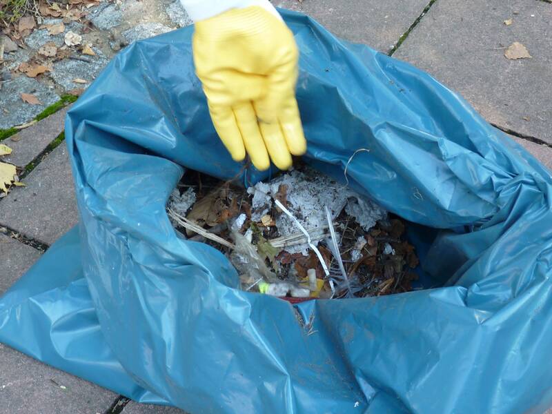 Eine Person mit gelbem Handschuh hält ihre Hand über einen offenen blauen Müllsack auf einer gepflasterten Fläche, in dem sich Unrat und Blätter befinden. (KI generiert)