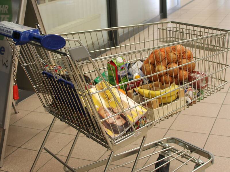 Ein Einkaufswagen voller Lebensmittel, einschließlich Bananen und Brötchen, steht in einem Flur neben einer Pflanzen und einer Werbetafel.