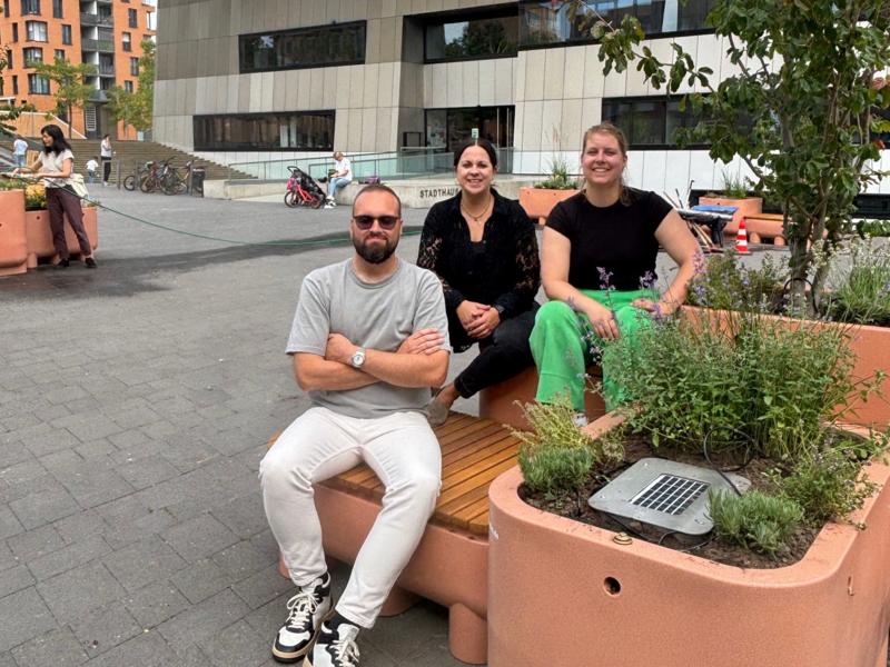 Drei Personen sitzen auf den Möbeln der Pop-Up-Aktion Schattenplatz vor dem Stadthaus im Scharnhauser Park, umgeben von Pflanzen und modernen Gebäuden.