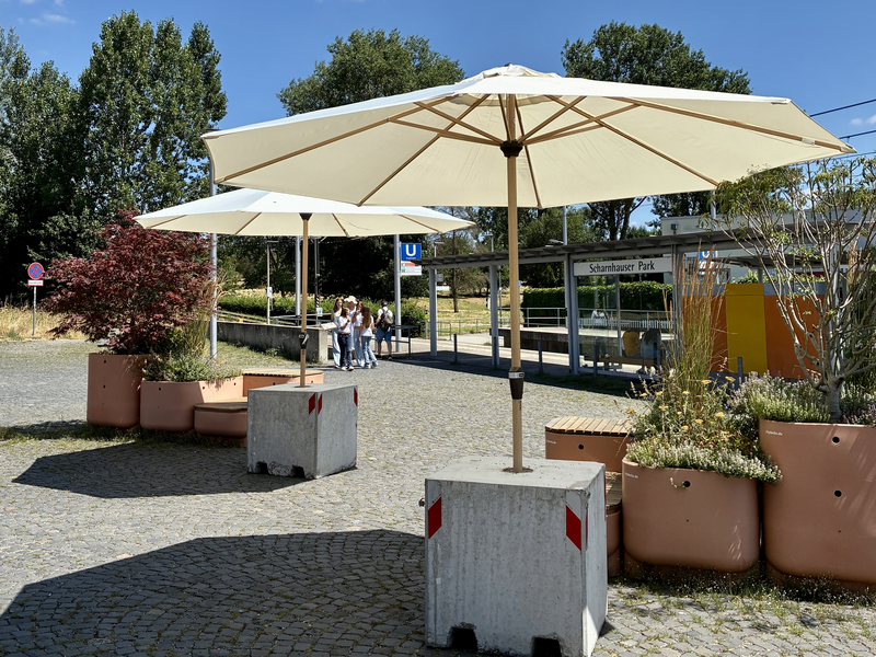Zwei große Sonnenschirme stehen auf einem gepflasterten Platz neben einer U-Bahn-Station, umgeben von Pflanzenkübeln und einigen Menschen im Hintergrund.