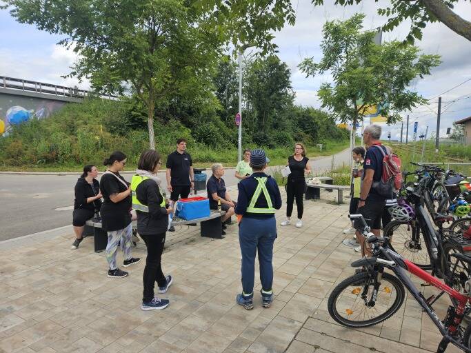 Eine Gruppe von Menschen steht auf einem gepflasterten Platz im Freien neben geparkten Fahrrädern und hört einer Person zu.