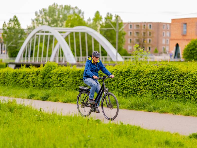 Eine Person fährt auf einem Fahrrad einen Weg entlang, vorbei an grünen Büschen, mit einer Brücke und mehreren Gebäuden im Hintergrund.