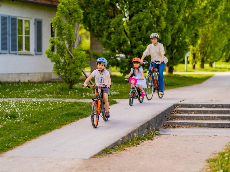 Ein Erwachsener und zwei Kinder fahren mit Fahrrädern auf einem Gehweg entlang einer grünen, baumbestandenen Straße.