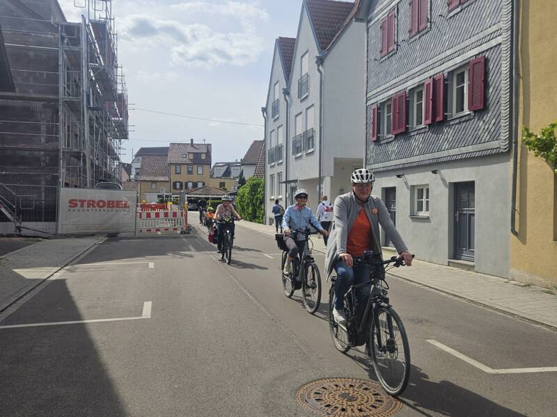 Das Bild zeigt eine Gruppe von Radfahrern, die auf einer ruhigen Straße in einem Wohngebiet unterwegs sind. Im Vordergrund ist ein Mann auf einem Fahrrad zu sehen, der einen Helm, eine graue Jacke und ein orangefarbenes Shirt trägt. Hinter ihm folgen mehrere weitere Radfahrer, alle ebenfalls mit Helmen. Rechts sieht man Häuser mit Giebeldächern und Fensterläden, links befindet sich eine Baustelle mit Gerüst und Absperrungen. Das Wetter ist sonnig mit blauem Himmel und einigen Wolken.