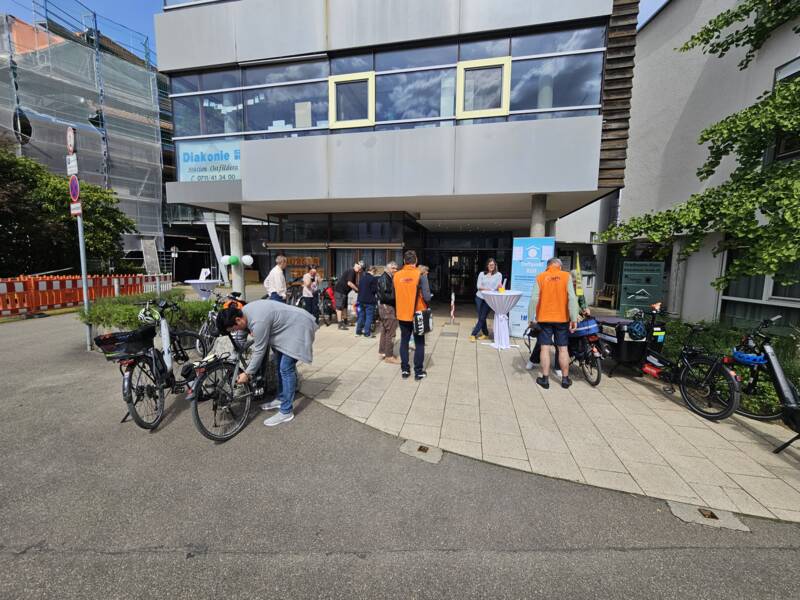  Auf dem Bild sieht man eine Gruppe von Menschen vor einem Gebäude mit großen Fenstern. Einige Personen tragen orangefarbene Westen mit dem Logo "adfc". Mehrere Fahrräder sind abgestellt. Im Hintergrund ist ein Schild mit dem Text "Treffpunkt RUIT" zu sehen. Das Wetter ist sonnig und die Szene wirkt freundlich und lebendig.