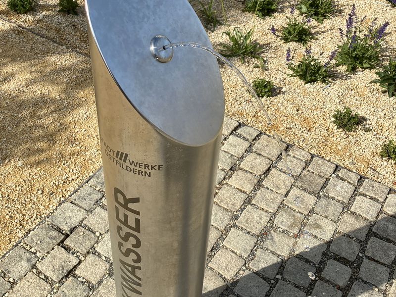 Eine Wasserstation aus Metall mit der Aufschrift "Trinkwasser" steht auf einem gepflasterten Gehweg, umgeben von einer mit Pflanzen bewachsenen Fläche.