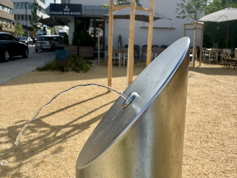 Eine moderne, schräg stehende Trinkwasserquelle mit der Aufschrift "Stadtwerke Ostfildern" sprudelt Wasser in einem gepflasterten Außenbereich, im Hintergrund ist ein Café zu sehen.