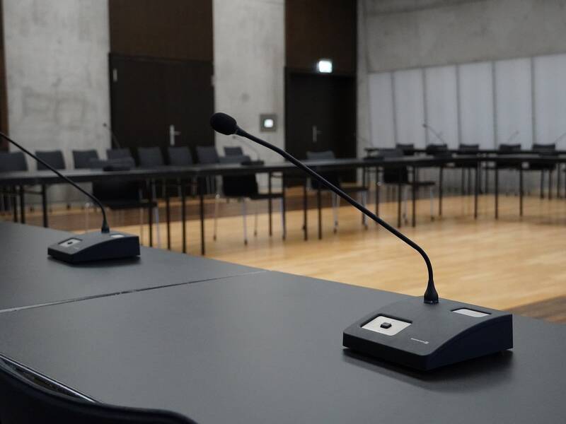 Ein Konferenzraum mit mehreren leeren Tischen und Stühlen sowie Mikrofonen auf den Tischoberflächen.