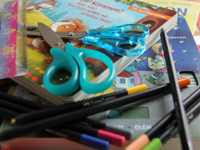 Ein Stapel Kinderbücher ist mit bunten Stiften, einer Schere und einem Lineal bedeckt.