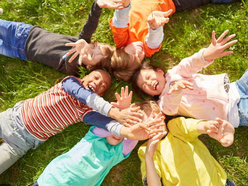 Eine Gruppe von lachenden Kindern liegt im Kreis auf einer grünen Wiese und streckt fröhlich die Hände in die Luft. KI-BildBeschreiber
