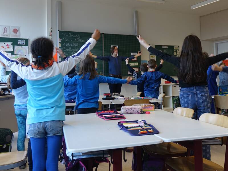 In einem Klassenzimmer stehen mehrere Kinder mit ausgebreiteten Armen um einen Tisch, während eine musikalische Lehrkraft ihnen etwas zeigt.